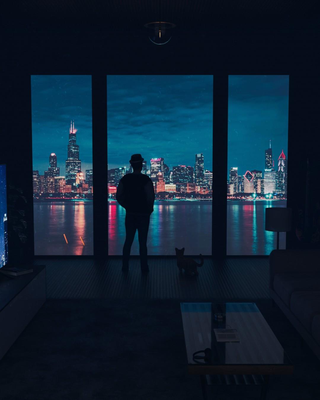 A man in a hat and his cat at a dark apartment's floor-to-ceiling windows, looking out at a city skyline lit up over water